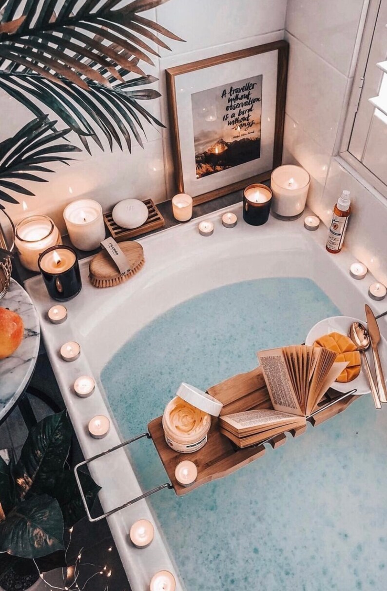 May include: A white bathtub filled with blue water, surrounded by lit candles and a wooden bath tray with a book, a bowl of fruit, and a jar of bath products. The text on the framed print reads "A traveler without observation is a bird without wings."