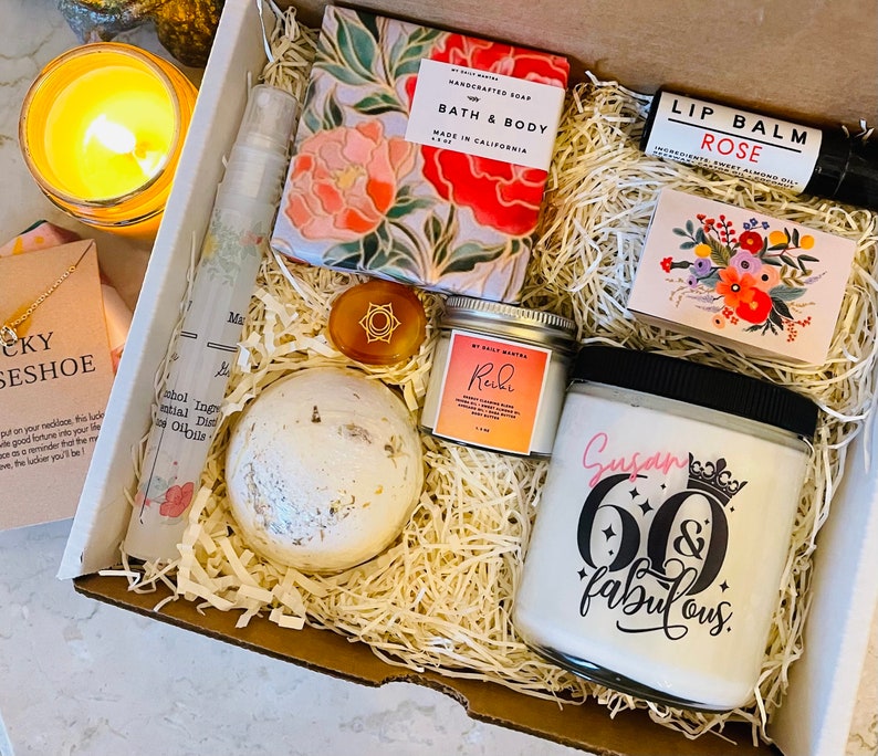 K&ouml;nnte beinhalten: Eine Geschenkbox gef&uuml;llt mit Artikeln f&uuml;r eine 60. Geburtstagsfeier. Die Box enth&auml;lt eine Seife f&uuml;r Bad und K&ouml;rper, einen Lippenbalsam, eine Badebombe, eine Kerze, ein kleines Glas Lotion und einen kleinen Dekorationsartikel. Die Kerze tr&auml;gt den Text "Susan 60 & fabulous".