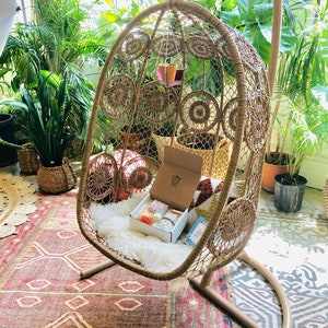 May include: A brown wicker hanging chair with a white faux fur cushion and a box of candles inside. The chair is hanging from a metal hook and is surrounded by plants.