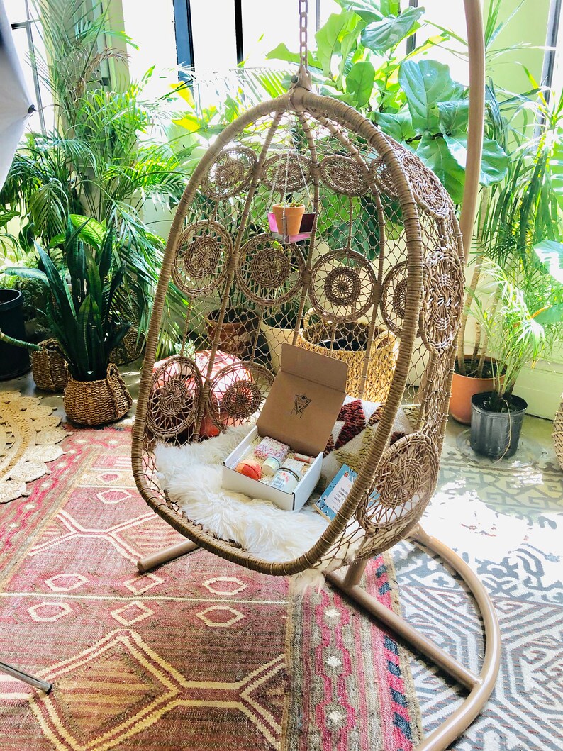 May include: A brown wicker hanging chair with a white faux fur cushion and a box of candles inside. The chair is hanging from a metal hook and is surrounded by plants.