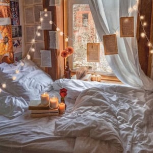 May include: A white bed with white bedding and pillows. There are string lights strung around the bed and a few candles on a stack of books. A small bouquet of red flowers is on the bed. The bed is in front of a window with white curtains and a view of a city street.