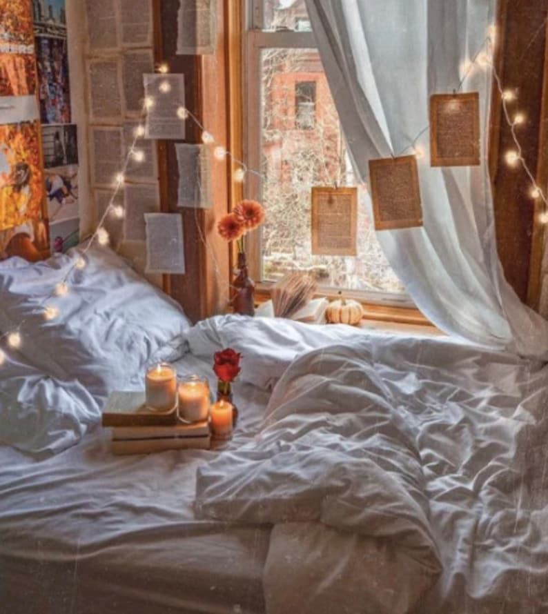 May include: A white bed with white bedding and pillows. There are string lights strung around the bed and a few candles on a stack of books. A small bouquet of red flowers is on the bed. The bed is in front of a window with white curtains and a view of a city street.