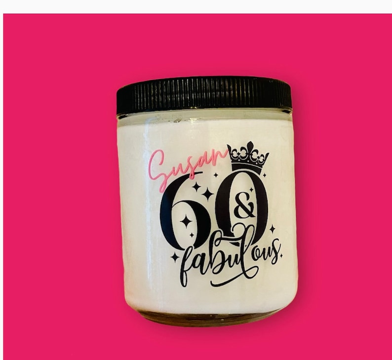 K&ouml;nnte beinhalten: Ein wei&szlig;es Kerzengef&auml;&szlig; mit schwarzem Deckel und einem schwarzen und rosafarbenen Design, auf dem "Susan 60 & fabulous" mit einer Krone &uuml;ber der 60 steht.