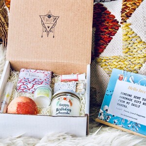 May include: A white gift box filled with a variety of self-care items, including a bath bomb, a candle, and a small jar of lotion. The box is decorated with a sticker that says "Happy Birthday Amanda". A blue card with the text "Hello lovely, Someone sent you a handmade gift box from my daily mantra." is visible in the background.