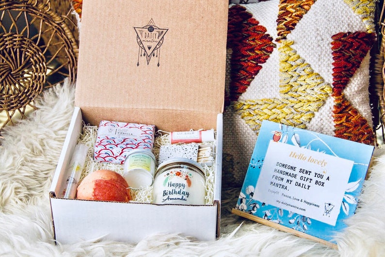 May include: A white gift box filled with a variety of self-care items, including a bath bomb, a candle, and a small jar of lotion. The box is decorated with a sticker that says "Happy Birthday Amanda". A blue card with the text "Hello lovely, Someone sent you a handmade gift box from my daily mantra." is visible in the background.