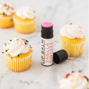 May include: A pink lip balm tube with a white label that reads "Lip Balm Vanilla Cupcake". The lip balm is sitting on a white marble surface with several cupcakes in the background.