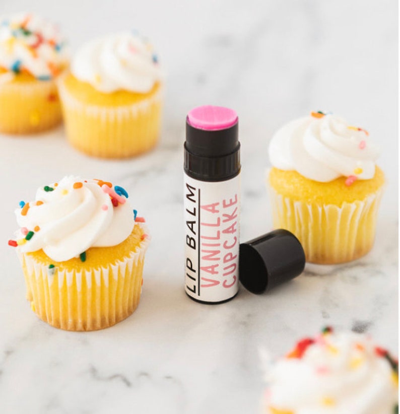 May include: A pink lip balm tube with a white label that reads "Lip Balm Vanilla Cupcake". The lip balm is sitting on a white marble surface with several cupcakes in the background.