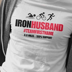 May include: White t-shirt with the text "IRONHUSBAND" in black and red, along with triathlon icons. Below the main text is "#TEAMFIRSTNAME" and "0.0 MILES - 100% SUPPORT". The shirt features a swimming, cycling, and running graphic.