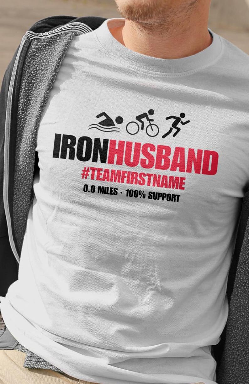 May include: White t-shirt with the text "IRONHUSBAND" in black and red, along with triathlon icons. Below the main text is "#TEAMFIRSTNAME" and "0.0 MILES - 100% SUPPORT". The shirt features a swimming, cycling, and running graphic.