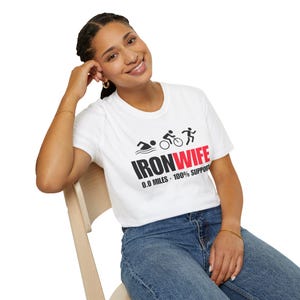 Può includere: Una t-shirt bianca a maniche corte con una grafica nera raffigurante un nuotatore, un ciclista e un corridore sopra la scritta IRONWIFE in nero e rosso, con il testo aggiuntivo 0.0 MILES - 100% SUPPORT stampato in nero sotto la grafica.