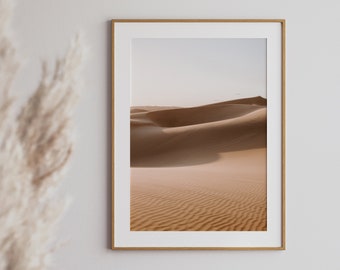 May include: A framed photograph of a desert landscape with rolling sand dunes. The sand is a light brown color and the sky is a pale blue.