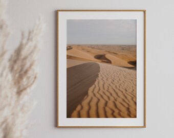 May include: A framed photograph of a desert landscape with rolling sand dunes. The sand is a warm beige color and the dunes are gently sloping. The sky is a light blue and there are no clouds visible.