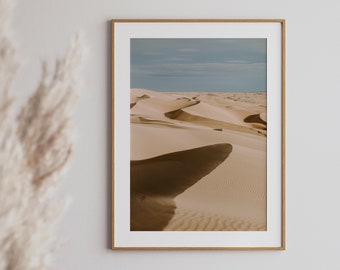 May include: A framed photograph of a desert landscape with rolling sand dunes. The sand is a light beige color and the sky is a light blue with some clouds.