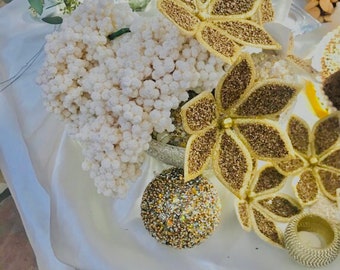 May include: A decorative arrangement featuring a white, textured pumpkin and gold glitter leaf accents. A round, beaded ornament and a silver napkin ring are also present. The arrangement is set on a white satin surface, with floral elements in the background.
