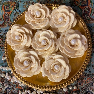 May include: A collection of seven tortilla-based flower arrangements, each with a pearl center, presented on a gold-colored, ornate tray. The tray is set against a backdrop of a patterned, teal-colored fabric, with scattered pearls.