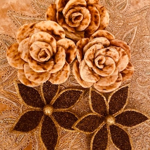 May include: Three brown, flower-shaped food arrangements sit atop a gold, patterned surface. Two decorative, dark brown flower shapes with gold glitter accents are below the food arrangements. The food flowers have a layered petal design.