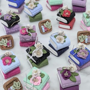 May include: A collection of miniature book-shaped earrings and square charms featuring stacked volumes in various colors. Each book is adorned with sculpted floral accents, including white, pink, and purple flowers with green leaves, and silver-toned hooks.