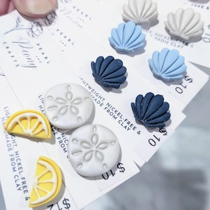 May include: A collection of stud earrings attached to white display cards labeled Pearl and Peony Crafts. The earrings feature clay shapes including light blue, dark blue, and white seashells, white sand dollars, and yellow lemon slices.