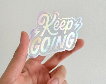 以下が含まれることがあります： 「Keep Going」の文字がスタイリッシュなフォントで書かれたホログラフィックステッカー。稲妻のグラフィックでアクセントが加えられています。ステッカーは真珠のような虹色の仕上がりで、さまざまな色を反射します。白い背景を背景に、手で持たれています。