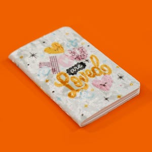Pode incluir: Um caderno de veludo branco com um design de glitter dourado e as palavras "You are loved" em letras de glitter dourado. O caderno tem um coração rosa com um rosto sorridente.