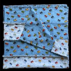 May include: A blue baby blanket with a patchwork design featuring a white background with a repeating pattern of colorful toy trains. The blanket has a frayed edge.