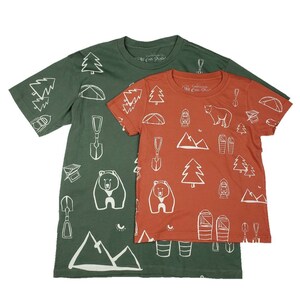 Peut inclure: Deux t-shirts sur le thème du camping. Le plus grand est vert avec des illustrations blanches d'ours, d'arbres, de tentes et d'autres équipements de camping. Le plus petit est orange avec les mêmes illustrations en blanc. Les deux t-shirts portent le texte "California All-en-Plein-Air" sur le devant.