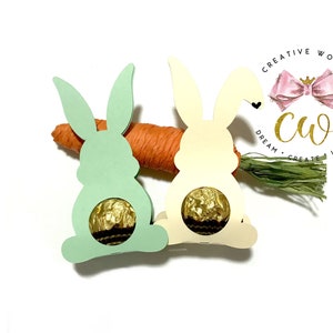 Op de afbeelding: Twee konijnenvormige kartonnen houders, een groene en een crèmekleurige, met een chocolade-ei in elk. De konijnen zitten op een wortel en gras. De tekst "Creative World" staat in een cirkel met een roze strik en de tekst "Dream Create Inspire" staat eronder.