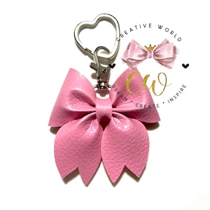 May include: A pink leather bow keychain with a silver heart-shaped clasp. The bow has a textured surface and is centered on a white background. The image also includes a logo with the words "CREATIVE WORLD" and "DREAM CREATE INSPIRE".