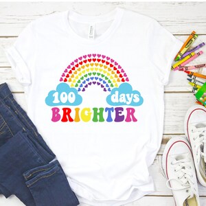 Puede incluir: Camiseta blanca con un dise&ntilde;o de arco&iacute;ris de corazones y el texto "100 d&iacute;as m&aacute;s brillantes" en letras de colores.