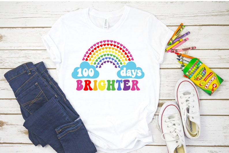Puede incluir: Camiseta blanca con un dise&ntilde;o de arco&iacute;ris de corazones y el texto "100 d&iacute;as m&aacute;s brillantes" en letras de colores.