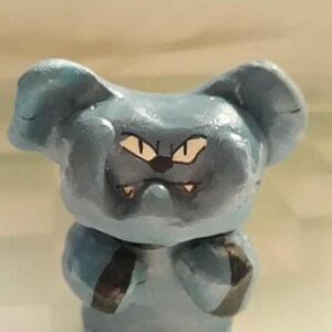 May include: A blue ceramic figurine of a bear with a grumpy expression. The bear has black eyes and a black nose. The bear is standing on its hind legs with its front paws raised.
