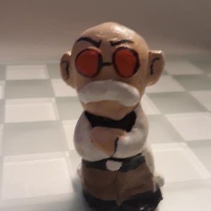 May include: A small ceramic figurine of an older person with a mustache and glasses. The figurine is wearing a white shirt and brown pants. The figurine has red eyes and a grumpy expression.