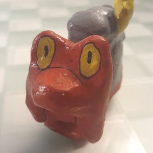 May include: A handmade, whimsical sculpture of a frog. The frog is painted in vibrant red with large yellow eyes outlined in black. A gray, textured form is behind the frog, with a yellow appendage extending upwards.