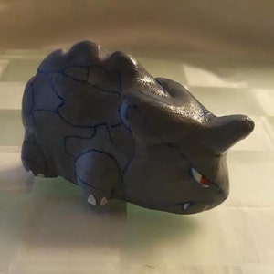 May include: A gray and blue ceramic figurine of a Pokemon character. The figurine is in a crouching position with its head down and its eyes closed. The figurine has a small horn on its head and a small tail.