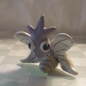 May include: A hand-crafted ceramic figurine of a purple and white butterfly with large eyes. The butterfly is sitting on a white surface.