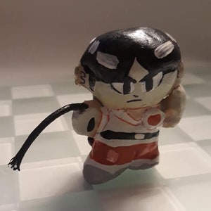 May include: A hand-painted figurine of a cartoon character with black hair, a white shirt, a red belt, and red shorts. The character is holding a black whip.