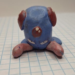 May include: A blue and red clay figurine of a Pokemon character with white spots. The figurine is sitting on a white gridded surface.