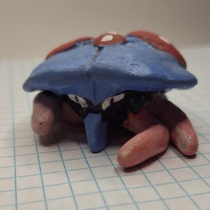 May include: A hand-painted ceramic figurine of a blue and red crab with white spots. The crab is in a crouching position with its claws tucked in.
