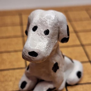 May include: A small, white ceramic figurine of a Dalmatian dog with black spots. The dog is sitting with its head tilted down and its eyes closed.