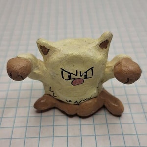 May include: A hand-painted ceramic figurine of a Pokemon character, a light brown and yellow Hitmonchan, with brown fists and a pink nose.