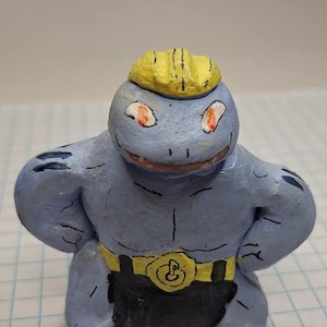 May include: A blue and yellow hand-painted clay figurine of a Pokemon character. The figurine has a yellow crown, red eyes, and a black belt with a gold buckle.