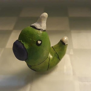 May include: A green and white hand-painted clay figurine of a cartoon caterpillar wearing a white hat. The caterpillar has a purple nose and black eyes.