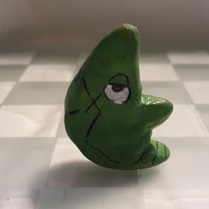 May include: A green ceramic crescent moon figurine with a white eye and black markings. The figurine is painted to look like a cartoon character.