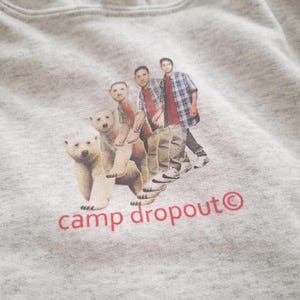 Puede incluir: Una sudadera gris con un estampado gráfico de cuatro personas y tres osos polares caminando. El texto "camp dropout" está impreso en rojo debajo del gráfico.