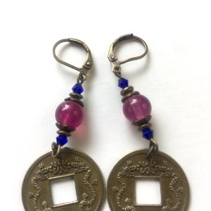 May include: A pair of gold-toned earrings with a pink and blue bead design. Each earring features a round coin with a square hole in the center and a dragon design around the edge.