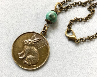 May include: A bronze necklace with a round pendant featuring a rabbit and Chinese characters. The pendant is attached to a chain with a turquoise bead.