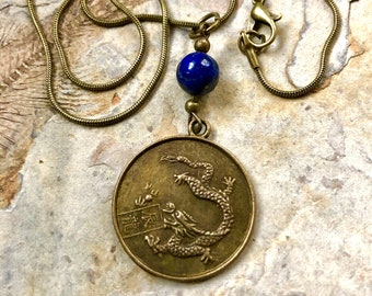 May include: A brass necklace with a round pendant featuring a dragon design and Chinese characters. The pendant is attached to a chain with a blue lapis lazuli bead.