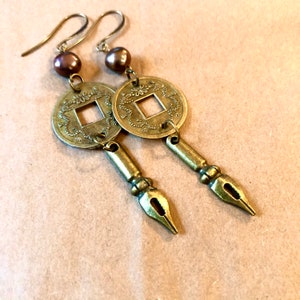 May include: A pair of gold-toned earrings featuring a coin-shaped charm and a pen-shaped charm. The coin charm has a square hole in the centre and intricate designs around the edge. The pen charm has a pointed tip and a decorative detail at the top.