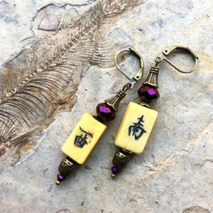 Op de afbeelding: Hangende oorbellen met crèmekleurige rechthoekige tegels met zwarte Chinese karakters. De oorbellen hebben bronzen metalen accenten, paarse kralen en oorhaken. Op een getextureerde, neutraal getinte achtergrond.