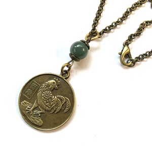 May include: A bronze necklace with a round pendant featuring a rooster design and Chinese characters. The pendant is attached to a chain with a green jade bead.